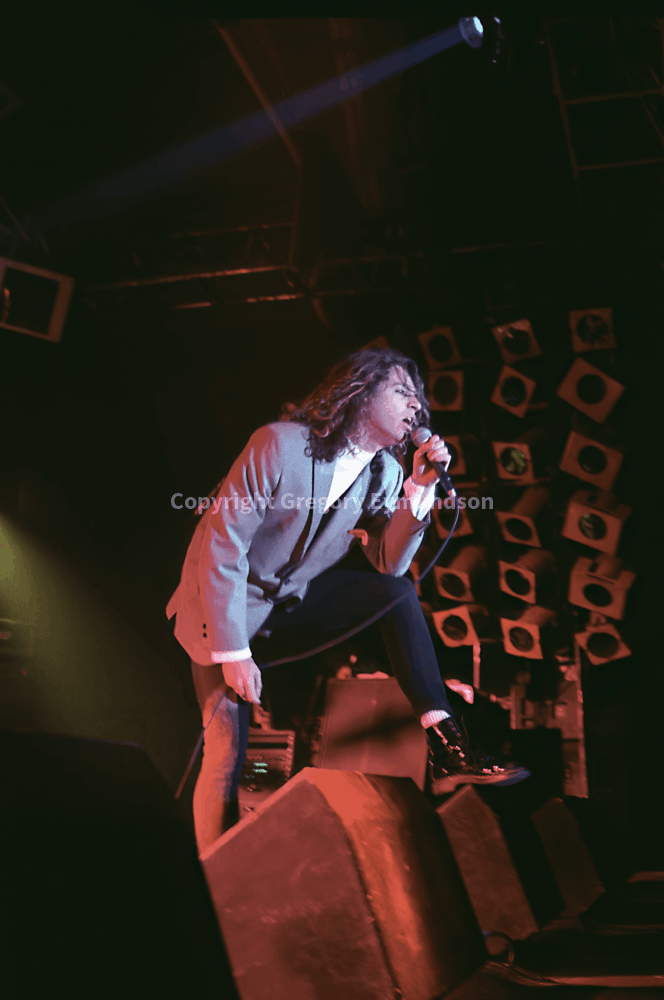 A performer passionately sings into a microphone, crouched on stage in a stylish outfit. Bright stage lighting highlights the energy of the live concert.