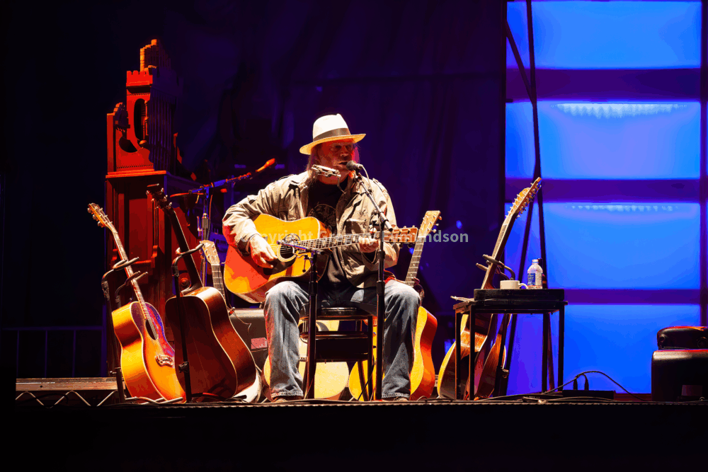 A musician in a hat performs on stage, seated with an acoustic guitar, surrounded by additional guitars and a glowing backdrop, showcasing a live music atmosphere.